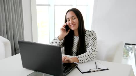 beautiful-secretary-talking-on-phone-in-white-office,-girl-manager-prints-text-on-laptop-at-desk,-employee-speaks-on-mobile