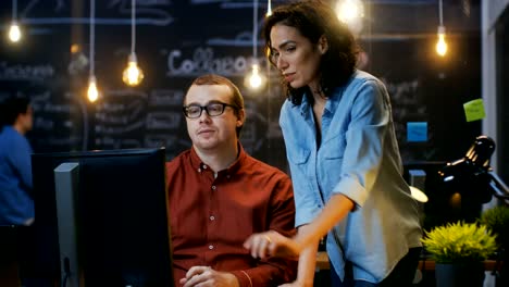 Männlicher-Büroangestellter-sitzt-an-seinem-Schreibtisch-und-Gespräche-mit-Kollegin,-She-Punkte-auf-seinem-Monitor.-Im-Hintergrund-kreativ-Büro.