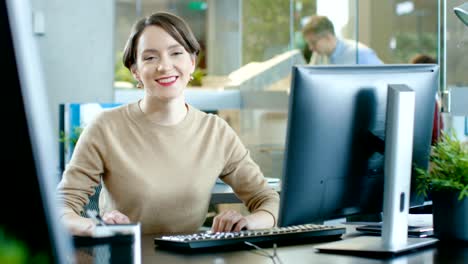 Hermosa-mujer-caucásica-joven-se-sienta-en-su-escritorio-y-trabaja-en-una-computadora-Personal.-En-el-fondo-oficina-ocupada-con-colegas-de-trabajo.