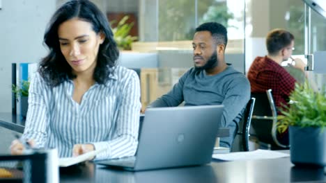 Oficina-Internacional-ocupada,-hermosa-mujer-hispana-trabajando-en-su-escritorio-en-un-portátil,-en-el-fondo-diverso-grupo-de-colegas-creativos-trabajando.