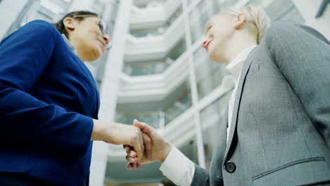 Low-angle-shot-of-businesswoman-shaking-hands-and-talking-with-female-business-colleague-in-suit-in-hall-of-modern-office-building