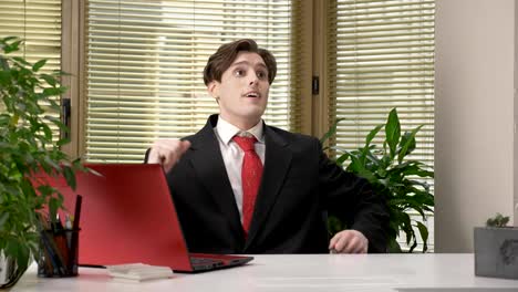 A-young-guy-in-a-suit-is-sitting-in-the-office-and-is-trying-to-throw-a-ball-for-American-football.-Comedy,-humor,-sport-concept.-60-fps