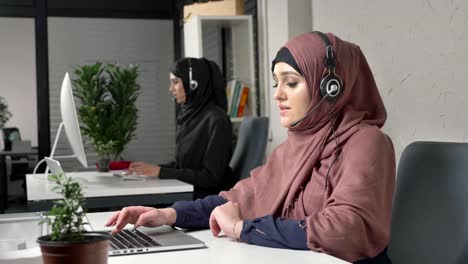 Una-joven-hermosa-en-un-hiyab-rosa-es-hablar-de-los-auriculares,-contestar-llamadas-en-call-center.-Mujeres-árabes-en-la-oficina.-60-fps