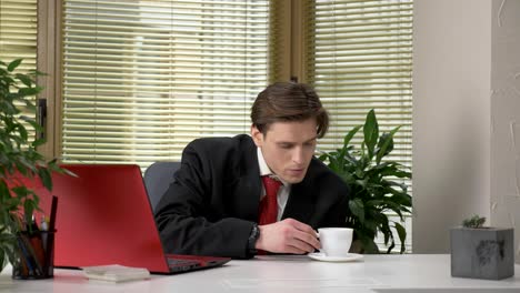 young-guy-in-a-suit-is-sitting-in-the-office,-working-on-a-laptop,-not-drinking-tasty-coffee,-making-funny-faces.-Work-in-office.-60-fps