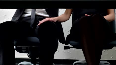 Shot-under-table,-young-woman-boss-sitting-beside-her-employee-and-touches-his-leg,-flirts,-harassment-concept-50-fps