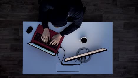 Young-man-in-black-hoodie-drinking-coffee-and-typing-on-laptop-during-night,-sitting-behind-white-desk,-top-shot