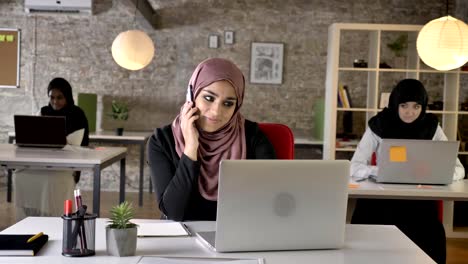 Tres-jóvenes-mujeres-musulmanes-en-hijab-sentada-y-trabajando-en-la-oficina-moderna,-hermosa-mujer-musulmana-hablando-por-teléfono,-sonriendo