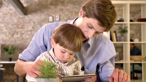 Joven-padre-tablet-viendo-con-su-pequeño-hijo,-niño-molesto-por-su-papá,-sentado-en-la-oficina-de-ladrillo-moderno