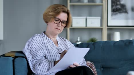 Old-Senior-Woman-Writing-on-Documents-while-Sitting-on-Sofa,-Paperwork