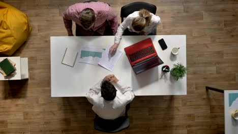 Team-of-three-young-business-people-coming-in-and-discussing-project,-topshot,-sitting-at-table-in-modern-office