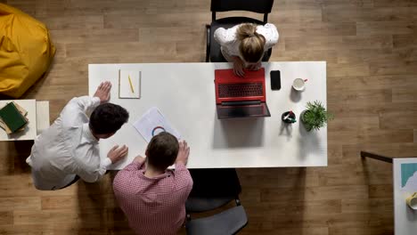 Dos-hombres-hablando-del-proyecto-cuando-mujer-trabajando-en-la-computadora-portátil-y-sentado-a-la-mesa-en-la-oficina-moderna,-topshot