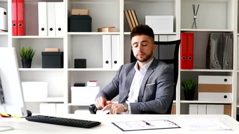 Junger-Geschäftsmann-in-graue-Jacke-sitzt-am-Tisch-in-weiß-Büro,-Blick-auf-Uhr-und-verlassen-auf-Stuhl