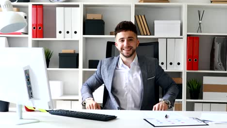 young-businessman-sitting-at-table-in-white-office,-smiling-and-showing-approving-hand-gesture