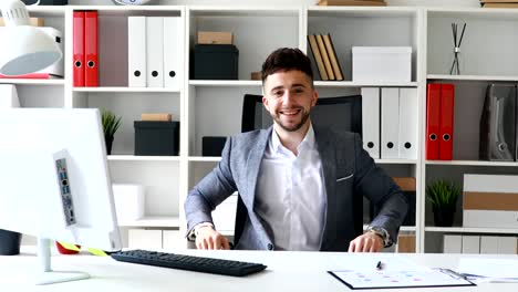 joven-hombre-de-negocios-sentado-a-la-mesa-en-la-oficina-de-blanco,-sonriendo-y-mostrando-aprobación-gesto-de-la-mano