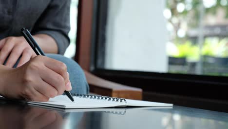 Mano-de-una-mujer-escribiendo-en-un-cuaderno-en-blanco-blanco-sobre-mesa