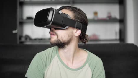 Joven-barbudo-a-hombre-hipster-usando-su-pantalla-del-auricular-VR-para-juego-de-realidad-virtual-o-ver-el-video-360-mientras-está-sentado-en-el-sofá-en-casa-en-la-sala-de-estar.-Tecnología-VR.