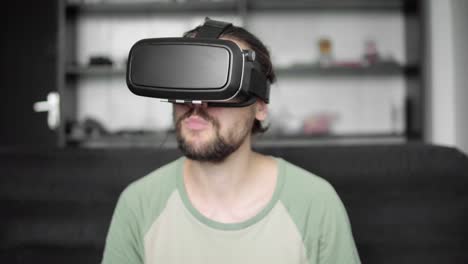 Young-bearded-hipster-man-using-his-VR-headset-display-for-watching-the-360-video-while-sitting-on-sofa-and-eating-cookies-at-home-in-the-living-room.-VR-Technology.