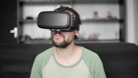 Junge-bärtige-Hipster-Mann-mit-seinem-VR-Kopfhörer-Display-zum-Betrachten-der-360-°-Video-auf-Sofa-sitzen-und-Essen-Cookies-zu-Hause-im-Wohnzimmer.-VR-Technologie.