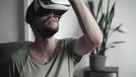 Young-bearded-hipster-man-using-his-VR-headset-display-for-virtual-reality-game-or-watching-the-360-video-and-trying-to-touch-to-something-he-see-while-sitting-on-sofa.-VR-Technology.