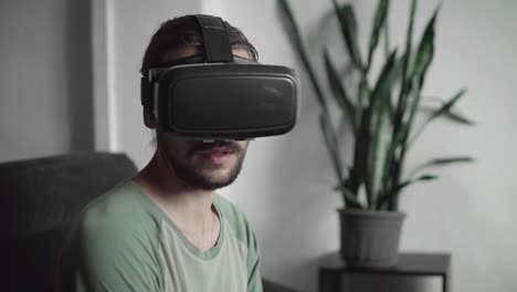 Young-bearded-hipster-man-using-his-VR-headset-display-for-virtual-reality-game-or-watching-the-360-video-and-trying-to-touch-to-something-he-see-while-sitting-on-sofa.-VR-Technology.