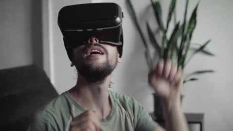 Young-bearded-hipster-man-using-his-VR-headset-display-for-virtual-reality-game-or-watching-the-360-video-and-trying-to-drive-away-what-he-sees-while-sitting-on-sofa.-VR-Technology.