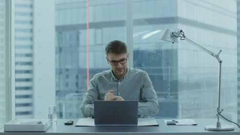 Handsome-Young-Businessman-Sitting-at-His-Desk-Using-Laptop-in-the-Office-Raises-Hands-and-Celebrates-Impressive-Fiscal-Results.-In-the-Background-Window-with-Cityscape-View.