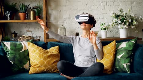 Zoom-in-of-smiling-young-lady-in-augmented-reality-glasses-moving-hands-and-turning-head-sitting-on-sofa-at-home-and-having-fun.-Entertainment-and-technology-concept.