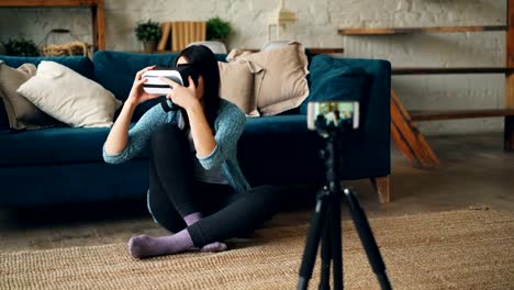 Attractive-lady-vlogger-is-using-smartphone-camera-to-record-video-about-virtual-reality-glasses.-Girl-is-holding-device,-showing-it-to-subscribers-and-talking.