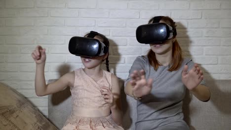 Zwei-junge-Mädchen-in-schwarzen-virtual-Reality-Brille-sitzen-und-virtuelle-Spiele-spielen.-Sie-winken-ihren-Händen,-virtuelle-Tasten-drücken-und-erkunden-Sie-den-umgebenden-Raum.