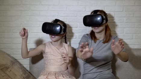 Two-young-girls-in-black-virtual-reality-glasses-sit-and-play-virtual-games.-They-wave-their-hands,-press-virtual-buttons-and-explore-the-surrounding-space.