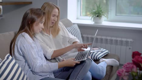 pair-of-friends-girls-are-sitting-on-a-couch-in-home-and-shopping-online,-discussing-and-holding-laptop-on-laps