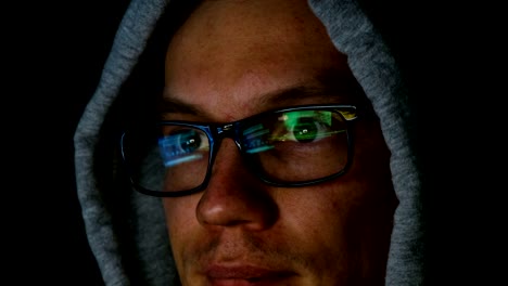 Hacker-in-glasses-and-a-hood-works-behind-a-computer-in-the-dark,-the-program-code-is-reflected-in-glasses-close-up,-4k