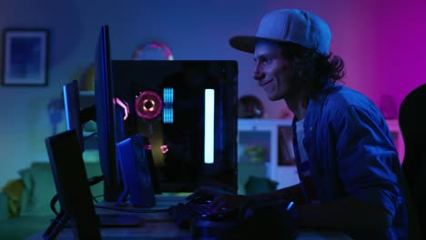 Emocionados-jugadores-jugando-y-ganando-en-First-Person-Shooter-juego-en-línea-de-vídeo-en-su-ordenador-Personal.-Sala-y-PC-tienen-colores-neón-Led-luces.-El-hombre-joven-lleva-una-gorra.-Agradable-noche-en-la-casa.