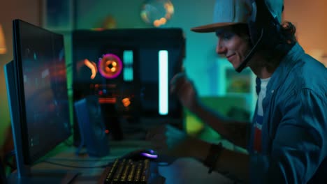 Gamer-Puts-His-Headset-with-a-Mic-On-and-Starts-Playing-Shooter-Online-Video-Game-on-His-Personal-Computer.-Room-and-PC-have-Colorful-Neon-Led-Lights.-Young-Man-is-Wearing-a-Cap.-Cozy-Evening-at-Home.