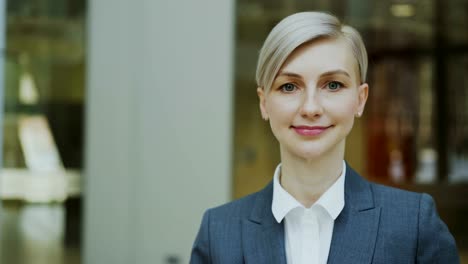 Close-up-portrait-of-successful-blonde-businesswoman-smiling-and-looking-into-camera-in-modern-office