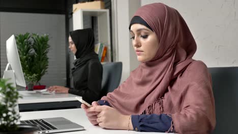 Young-beautiful-girl-in-pink-hijab-sits-in-office-and-uses-smartphone.-Girl-in-black-hijab-in-the-background.-Arab-women-in-the-office.-60-fps