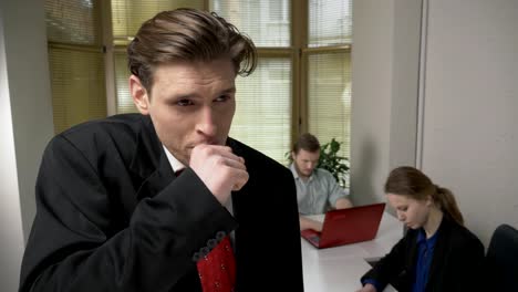 Young-man-in-a-suit-is-coughing,-sick,-a-disease-concept.-Man-and-the-girl-in-the-background-are-working-in-the-office.-60-fps