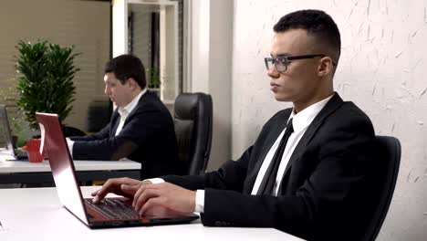 Joven-africana-hombre-de-negocios-acertado-con-gafas-está-sentado-en-la-oficina-trabajando-en-un-portátil,-saca-sus-gafas,-mira-a-la-cámara,-sonríe-y-sacude-su-cabeza,-caucásico-hombre-en-un-traje-en-el-fondo.-60-fps