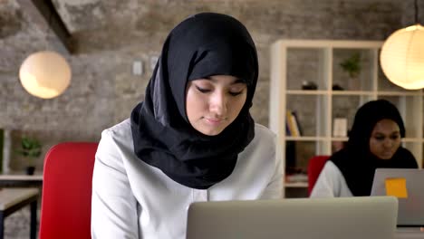 Retratos-de-mujeres-jóvenes-musulmanas-en-hijab-trabajando-y-mirando-en-la-cámara,-sonriendo,-dos-mujeres-sentadas-en-la-oficina-moderna