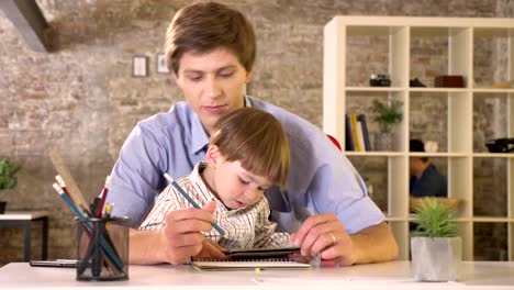 Young-father-holding-his-little-son-and-writing-on-paper,-kid-holding-tablet,-sitting-in-modern-office