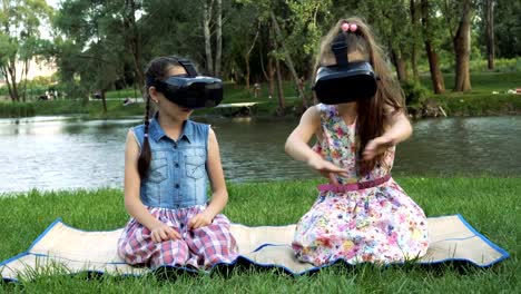 Zwei-kleine-Mädchen-spielen-virtuelle-Spiele-im-Park-am-Fluss-Bank.-Sie-sitzen-in-virtual-Reality-Brille,-schauen-Sie-sich-um-und-winken-ihren-Händen.