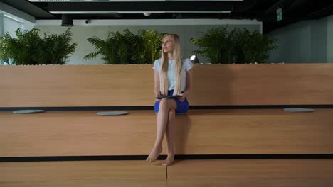 Relaxing-woman-with-tablet-in-office.-Young-elegant-woman-sitting-barefoot-on-wooden-tribune-in-modern-office-using-tablet-and-taking-rest