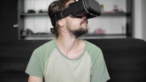 Junge-bärtige-Hipster-Mann-mit-seinem-VR-Kopfhörer-Anzeige-mit-Kopfhörern-für-virtual-Reality-Spiel-oder-das-360-Viedeo-beim-Sitzen-auf-dem-Sofa-zu-Hause-im-Wohnzimmer.-VR-Technologie.