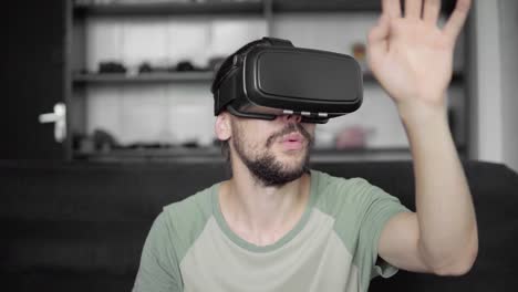 Young-bearded-hipster-man-using-his-VR-headset-display-for-virtual-reality-game-or-watching-the-360-video-and-trying-to-touch-to-something-he-see-while-sitting-on-sofa.-VR-Technology.