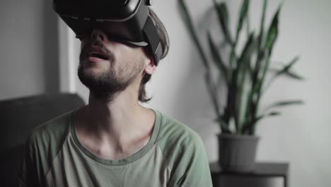 Joven-barbudo-a-hombre-hipster-usando-su-pantalla-del-auricular-VR-para-juego-de-realidad-virtual-o-ver-el-video-360-mientras-está-sentado-en-el-sofá-en-casa-en-la-sala-de-estar.-Tecnología-VR.