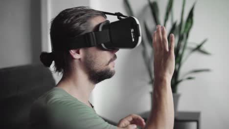 Young-bearded-hipster-man-using-his-VR-headset-display-for-virtual-reality-game-or-watching-the-360-video-and-trying-to-touch-to-something-he-see-and-catch-while-sitting-on-sofa.-VR-Technology.