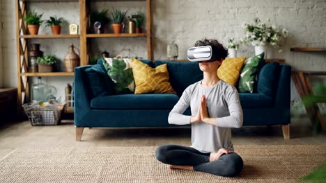 Slim-young-lady-in-artificial-reality-glasses-is-meditating-at-home-in-lotus-pose-with-hands-in-namaste-enjoying-simulation-and-relaxing.-Modern-technology-and-experience-concept.