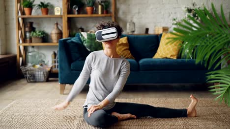 Smiling-girl-in-virtual-reality-glasses-is-training-sitting-on-floor-at-home-stretching-legs-and-body-and-enjoying-ar.-Healthy-lifestyle-and-vr-simulation-concept.