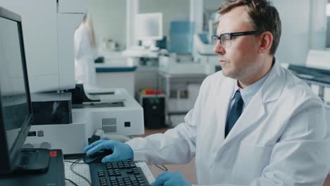 Male-Research-Scientist-Sits-at-His-Workplace-in-Laboratory,-Uses-Personal-Computer.-I-the-Background-Genetics,-Pharmaceutical-Research-Centre-with-Innovative-Equipment.