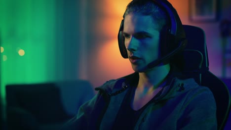 Gamer-in-Headset-with-a-Mic-Playing-and-Winning-in-Online-Video-Game-on-His-Personal-Computer.-He-Gives-Commands-to-Other-Players.-Room-and-PC-have-Colorful-Neon-Led-Lights.-Cozy-Evening-at-Home.